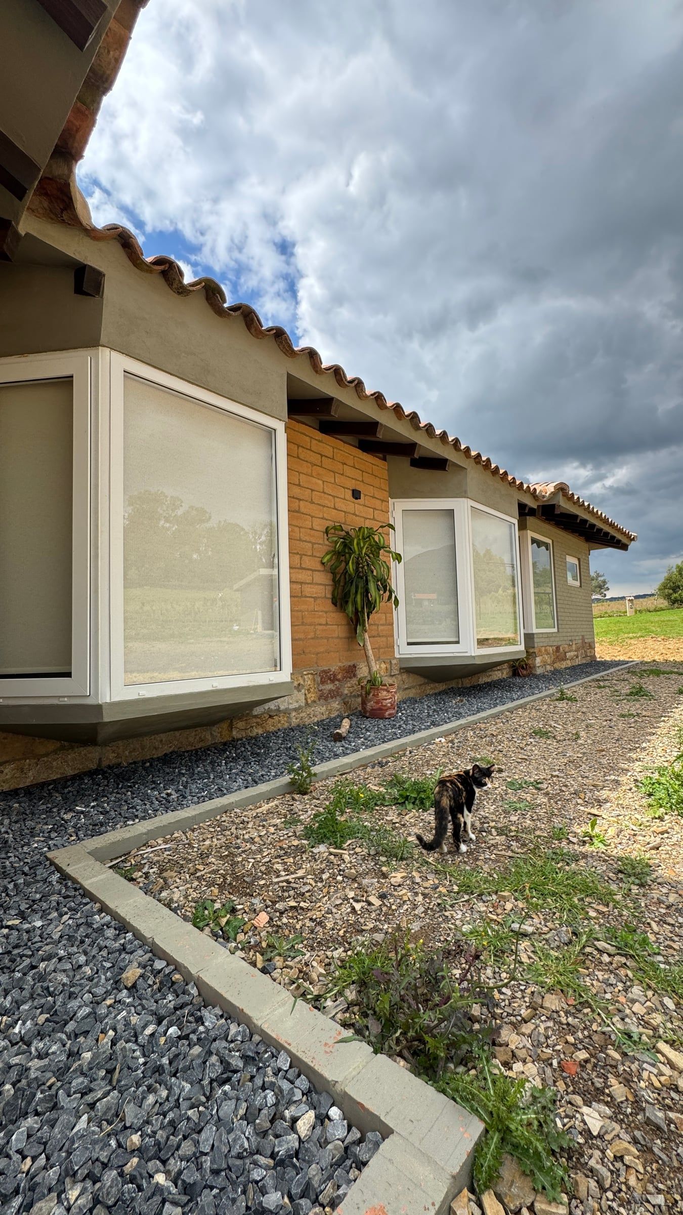 Fachada con ventana mirador, muro de tierra y alero de teja sobre un borde de gravilla