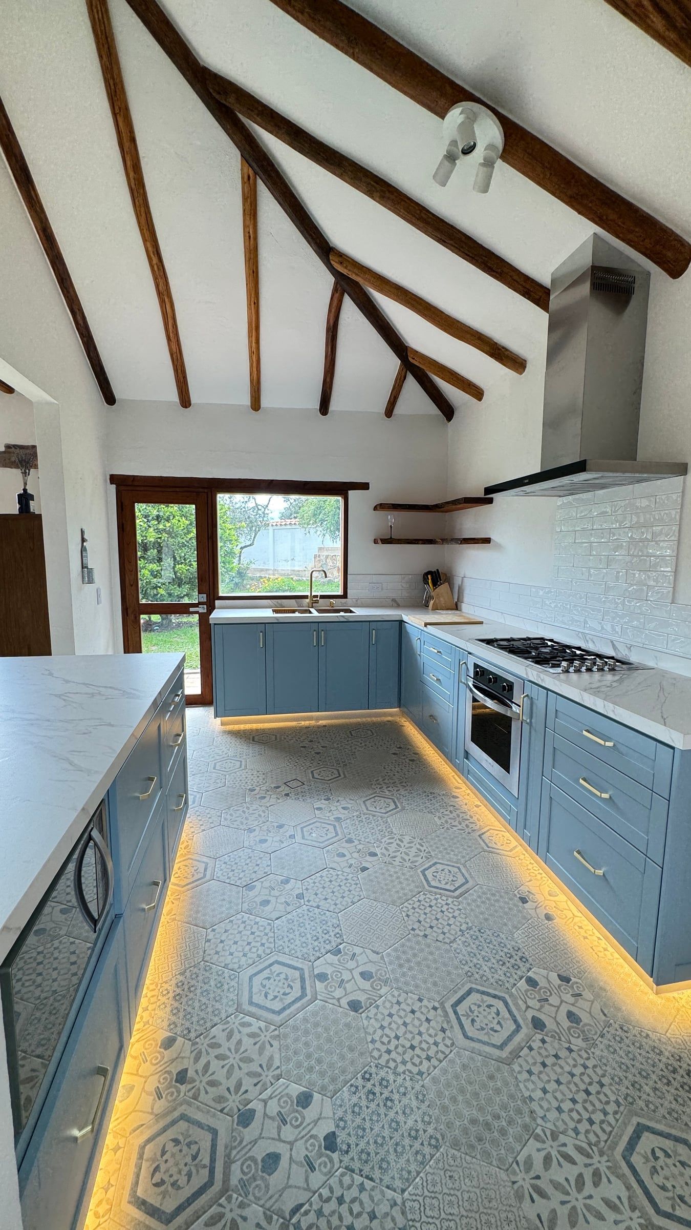 Casa Nido Verde después de la remodelación: cocina con muebles azules, mesón claro, piso de baldosa geométrica e iluminación bajo los muebles, bajo la misma cubierta de vigas